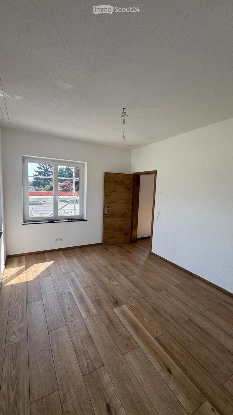 Empty room with wooden flooring, white walls, and a large window with a view of the outside. A wooden door is visible on the right side.