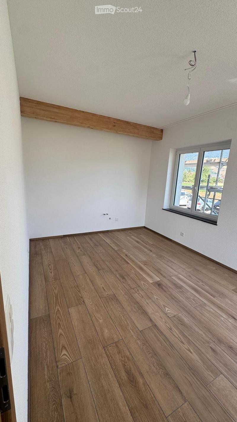 Empty room with wooden flooring, white walls, and a large window with a view of the outside. The room has a simple design with a wooden trim along the ceiling and a light switch visible on the wall.