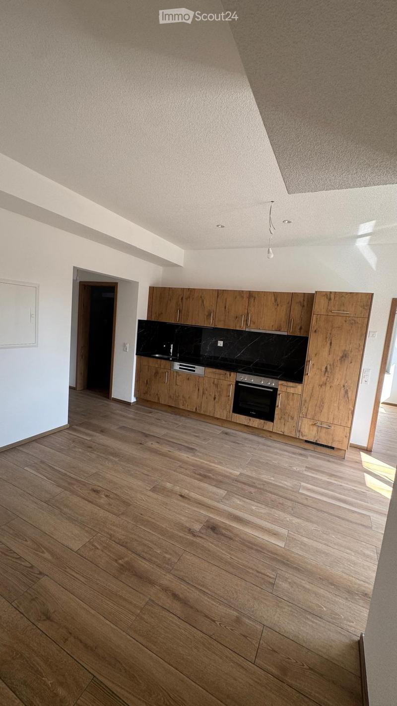 Modern kitchen with wooden cabinets, black countertops, and a built-in oven. The floor is made of light wooden planks, and the space is well-lit with recessed ceiling lights.