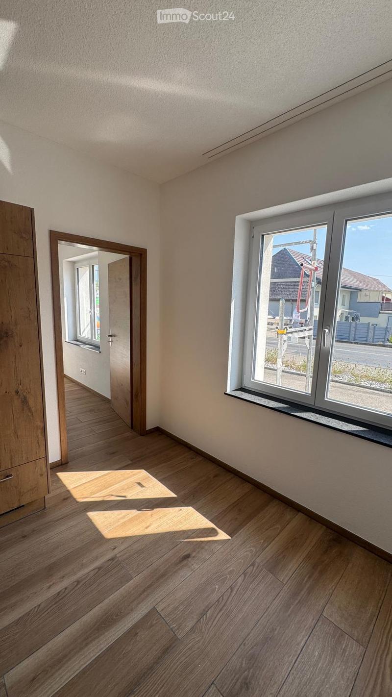 Empty room with wooden flooring, white walls, and large windows. The room has a door leading to another space and is well-lit with natural light.