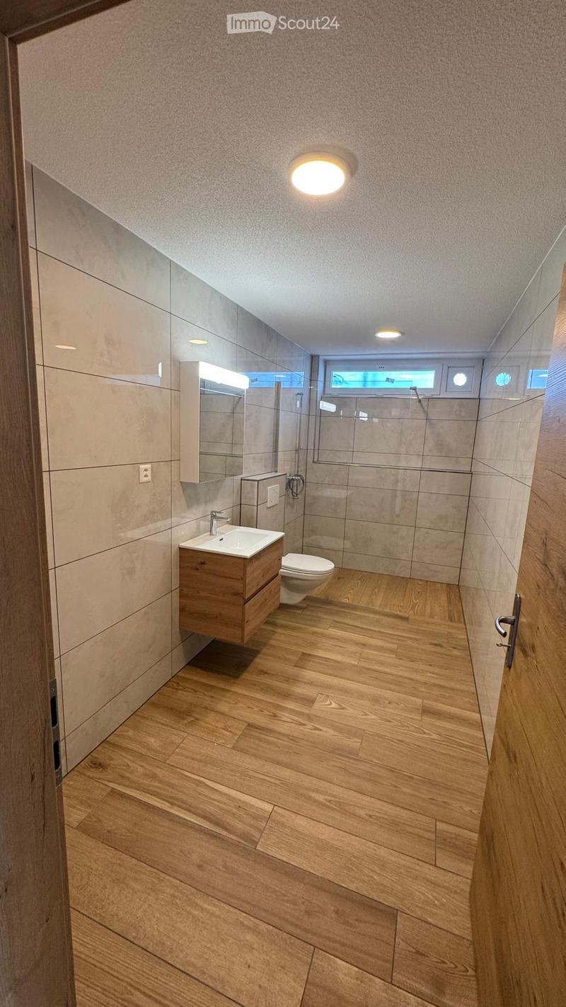 Modern bathroom with wooden flooring, a white sink with a wooden cabinet, a toilet, a large mirror, and a glass-enclosed shower area.