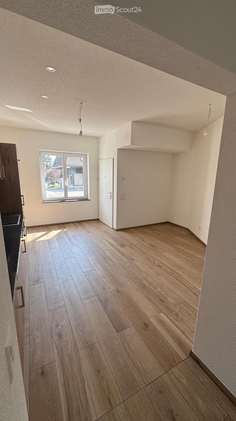 Empty room with wooden flooring, white walls, and a large window letting in natural light. The room features recessed ceiling lights and a door leading to another area.