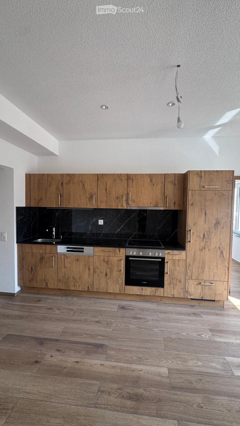 Modern kitchen with wooden cabinets, black countertops, and stainless steel appliances including an oven and dishwasher. The floor is made of light wood.