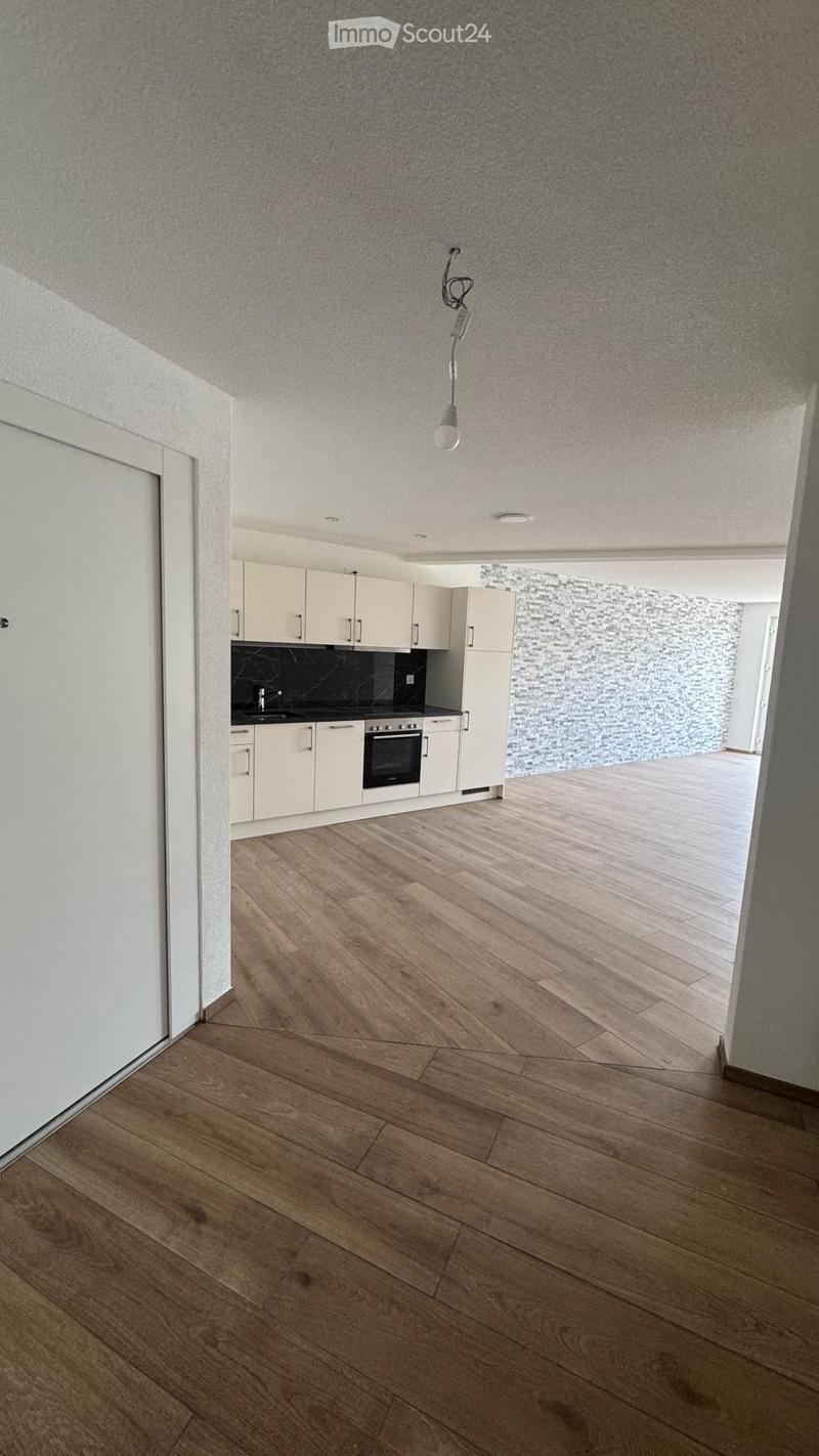 Empty room with wooden flooring, white walls, and a small kitchen area with cabinets and appliances.