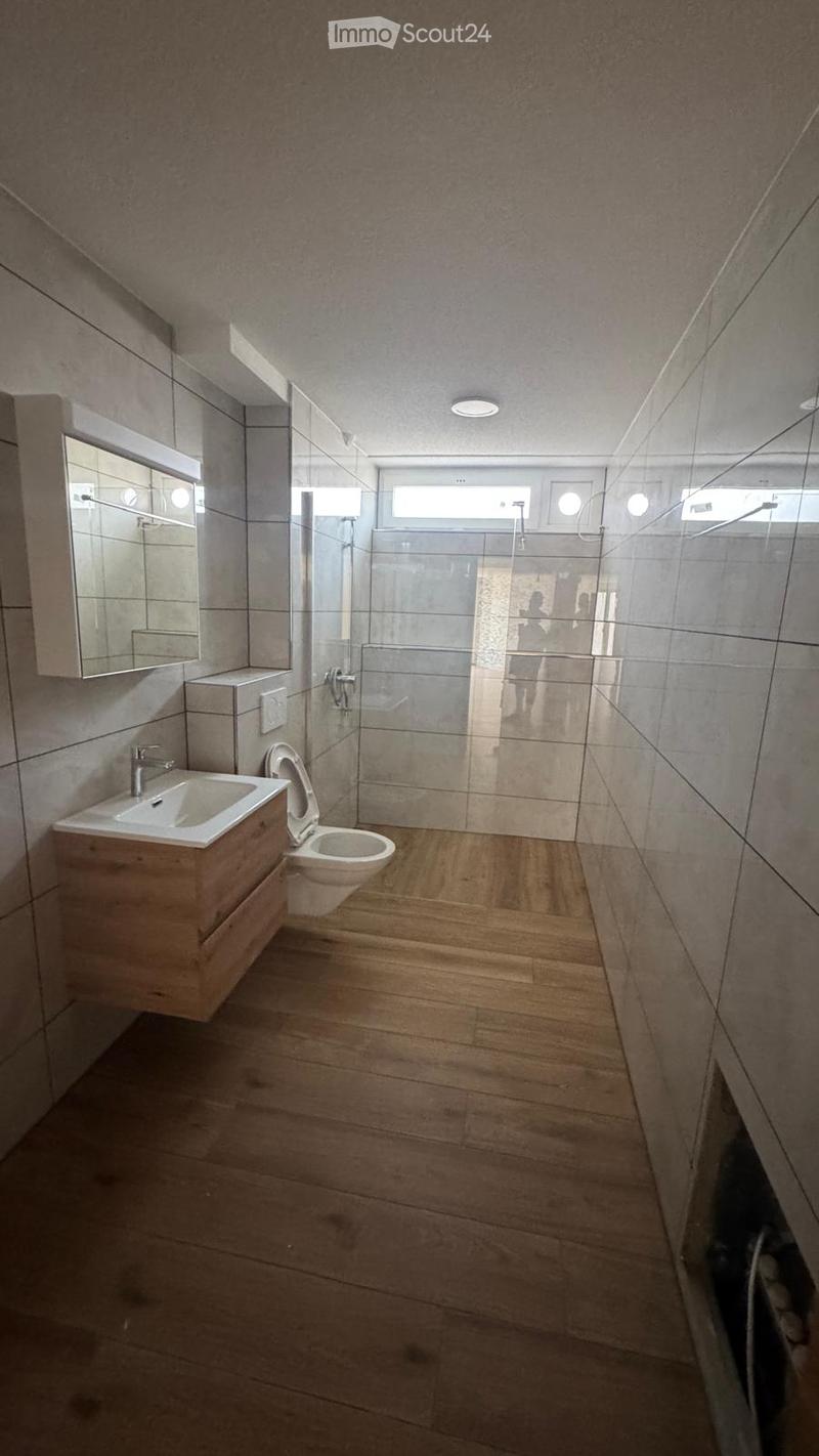 Modern bathroom with wooden flooring, a sink with a faucet, a toilet, a mirror cabinet, and a glass-enclosed shower area.