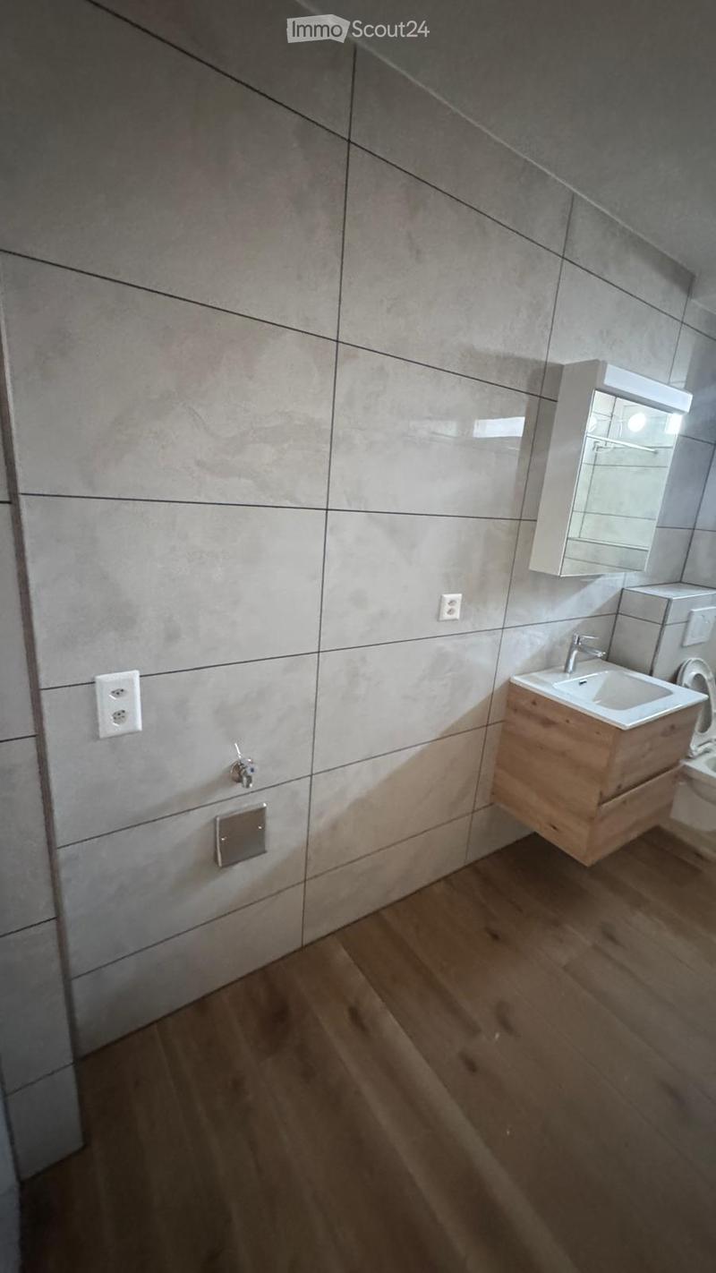Modern bathroom with light grey tiled walls, wooden floor, white sink with wooden cabinet, mirror, and toilet.