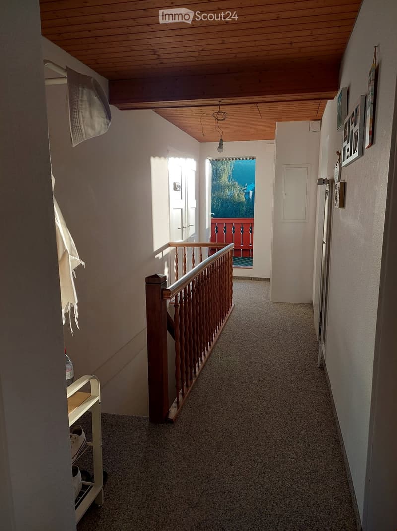 Hallway with wooden railing, carpeted floor, and a view of a balcony with a red railing and outdoor scenery.