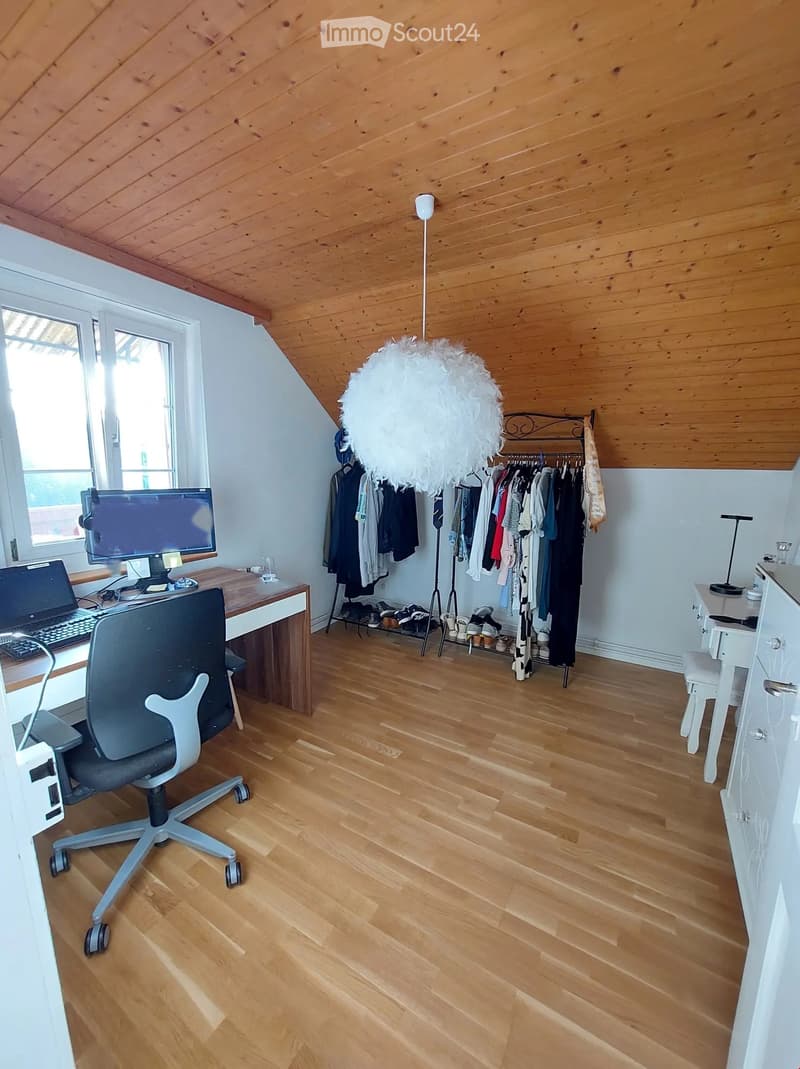 Bright, wooden-floored room with a slanted wooden ceiling, featuring a desk with a laptop, screen, and keyboard, a chair, a wardrobe with hanging clothes and shoes, and a decorative hanging lamp.
