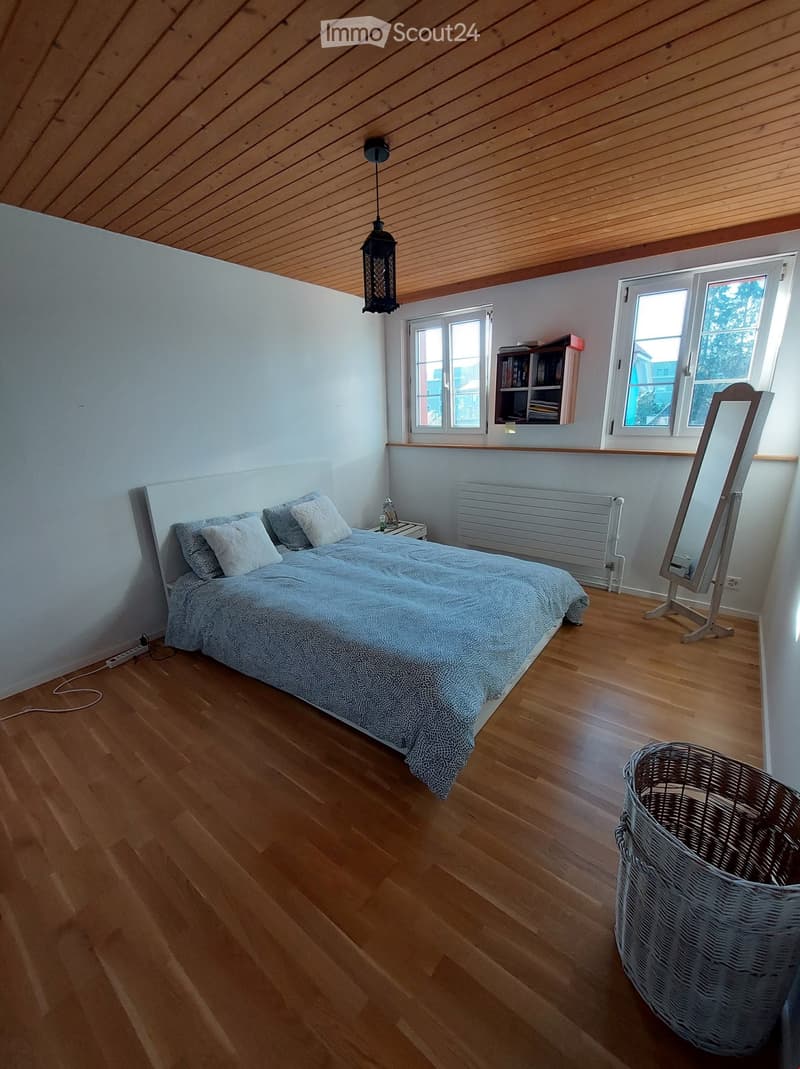 Bright bedroom with wooden flooring, a large bed with pillows and a patterned duvet, a nightstand with a lamp, a mirror on a stand, a radiator, and a bookshelf with books.