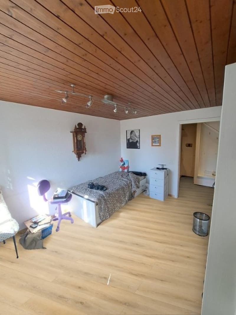 Cozy bedroom with wooden flooring and ceiling, featuring a bed with a patterned bedspread, a purple chair, a desk with a lamp, a wooden cabinet, and a wall-mounted clock.
