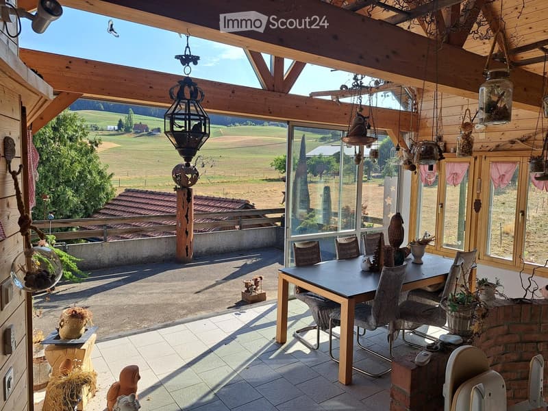 Bright, wooden-framed balcony with large windows offering a scenic countryside view. Features a dining table with chairs, hanging decorations, and potted plants.