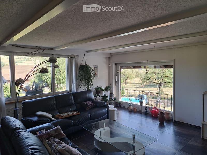 Modern living room with large windows, a black leather sofa, a glass coffee table, a floor lamp, and several potted plants. The room has a view of a garden and a swimming pool through the windows.