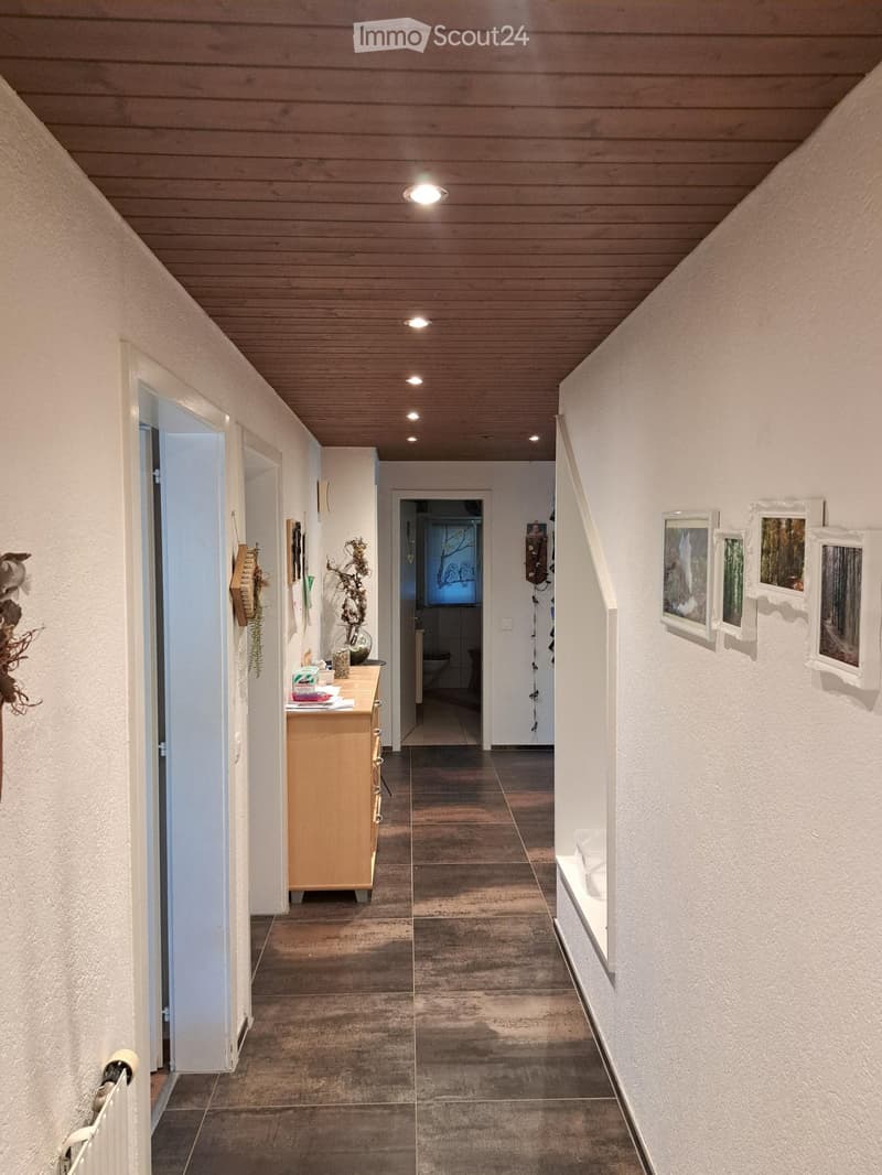 A modern hallway with white walls, a wooden ceiling, and recessed lighting. Features a small wooden console table with a vase and decorative items, framed artwork on the walls, and a visible bathroom entrance.