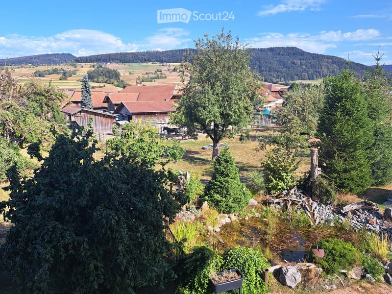 A rural garden area with trees, shrubs, a small water feature, and a view of a valley with houses and hills in the background.