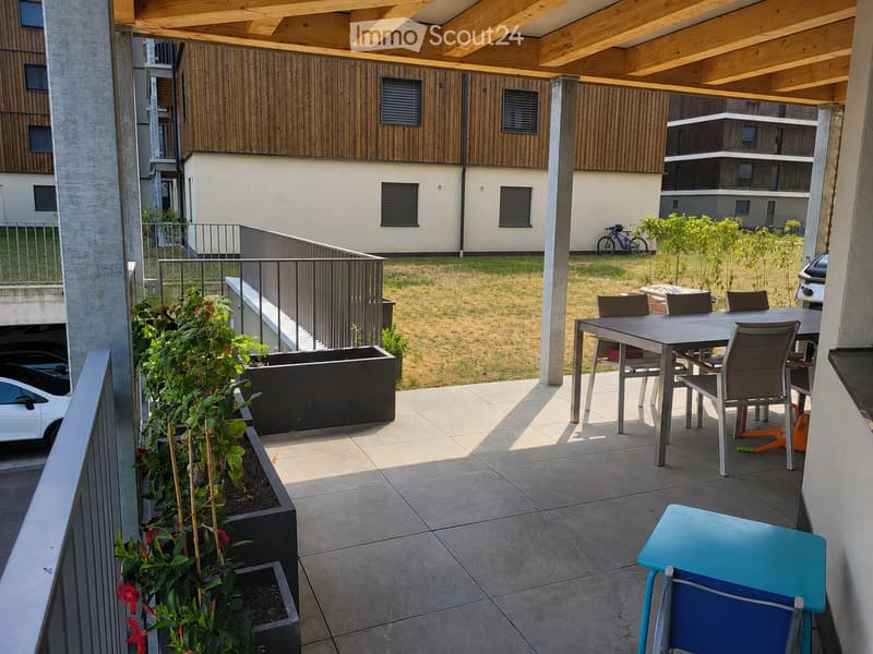 Balcony with a modern table and chairs, surrounded by greenery and overlooking a grassy area with buildings in the background.