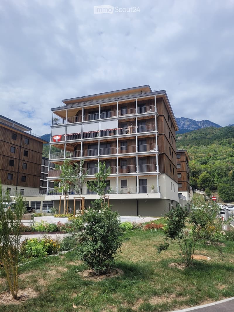 Modern multi-story building with wooden balconies and a Swiss flag, surrounded by greenery and mountains.