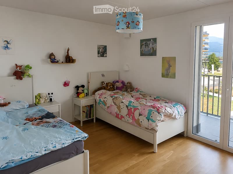 Bright, child-friendly bedroom with two single beds, colorful bedding, nightstand, toys, and artwork on the walls. Large window offers a scenic view.