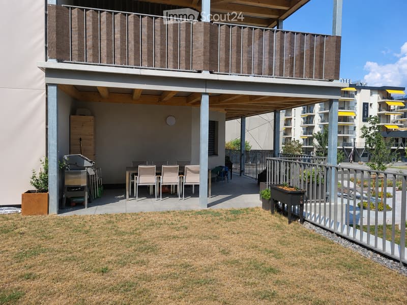 Balcony with a covered seating area, a grill, a table with chairs, and potted plants. The balcony is part of a modern residential complex with contemporary design elements.