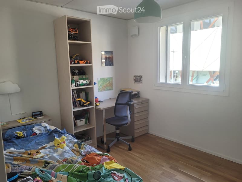 A well-lit bedroom with a bed, desk, chair, and bookshelf filled with books and toys. The room features wooden flooring and large windows.