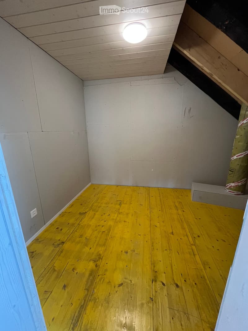 Empty room with yellow wooden flooring, white walls, and a slanted ceiling with exposed beams. A single light fixture is mounted on the ceiling.