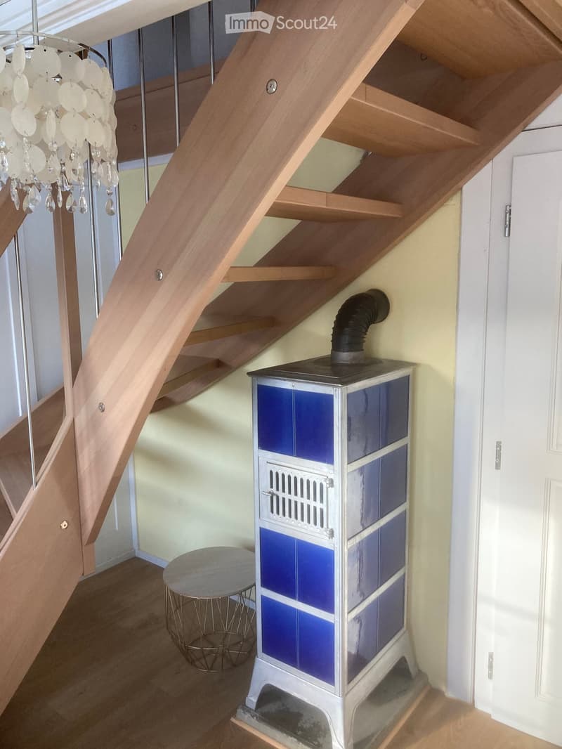Wooden staircase with a chandelier, a small round table, and a blue-tiled wood-burning stove in the corner.