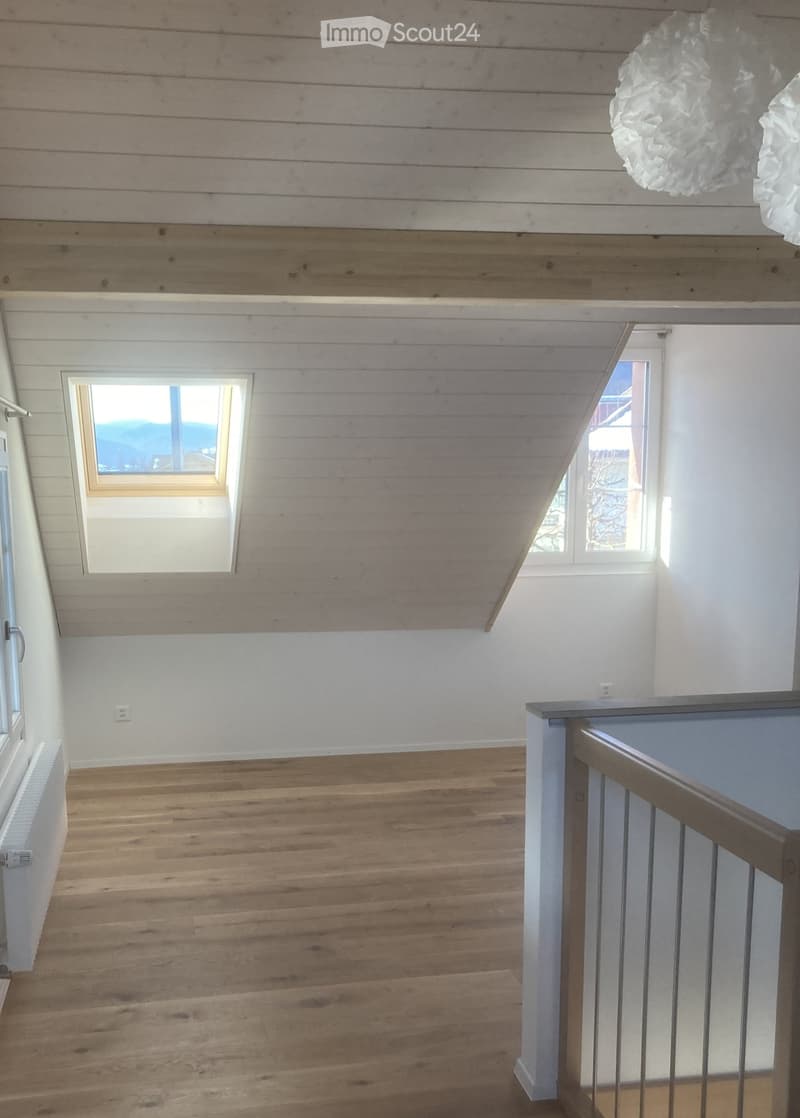 Empty room with wooden flooring, white walls, and exposed wooden ceiling beams. Features a staircase railing and a small window with a scenic view.