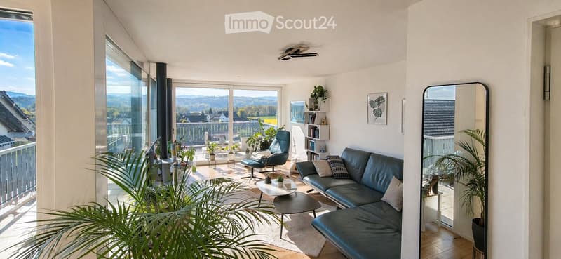 Bright living room with large windows and a view, featuring a dark blue sofa, coffee table, armchair, and decorative plants.