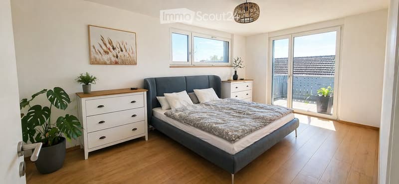 Modern bedroom with a grey upholstered bed, white pillows, a wooden nightstand, a drawer unit, potted plants, and a large window with a view to a balcony.