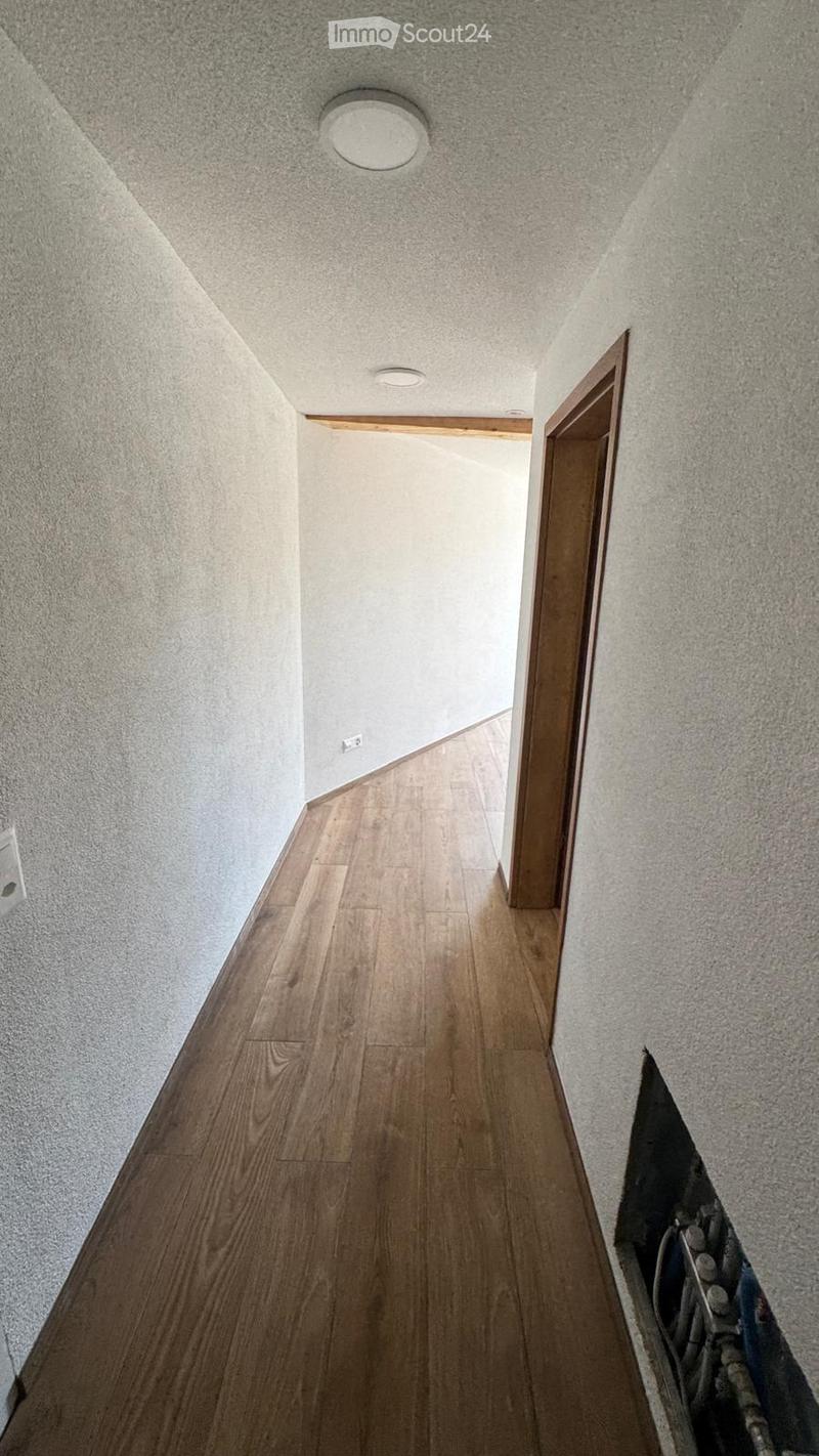 A narrow hallway with wooden flooring, white walls, and recessed lighting. A wooden door is visible on the right side, and there is a small opening showing pipes and wiring on the lower right corner.