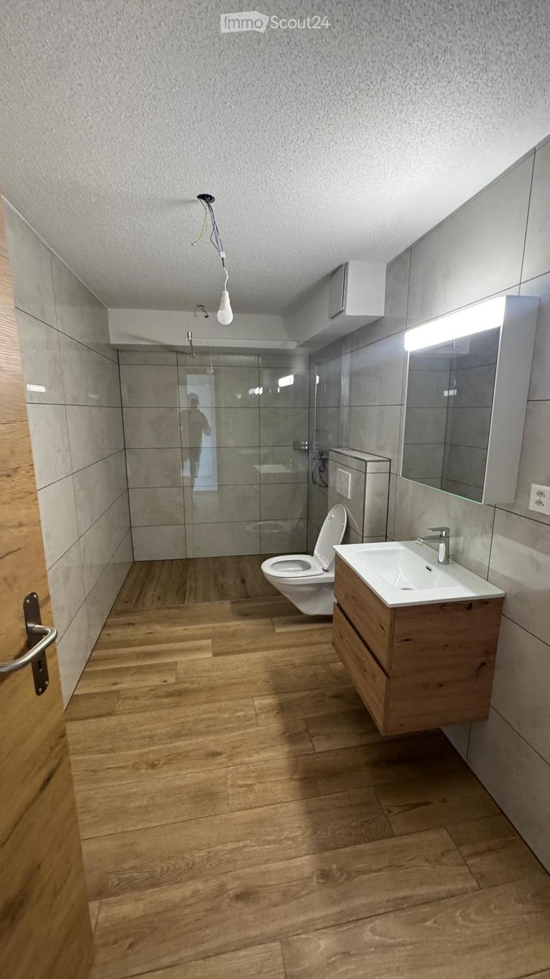 Modern bathroom with wooden flooring, a toilet, a sink with a wooden cabinet, a large mirror, and a glass-enclosed shower area.