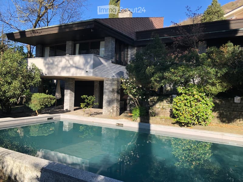 Modern two-story house with a flat roof, large windows, and a swimming pool in the foreground. The house features a raised ground floor and is surrounded by lush greenery and trees.