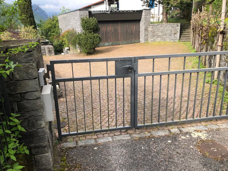 A metal gate with a lock, leading to a paved area with a stone wall and a small garden, with a house and garage visible in the background.