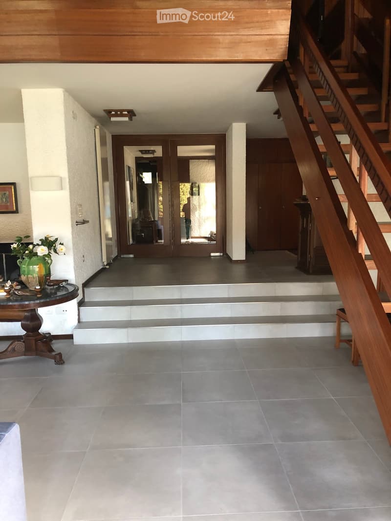 Spacious hallway with tiled flooring, wooden staircase, and a round wooden table with a vase of white flowers on the left side.