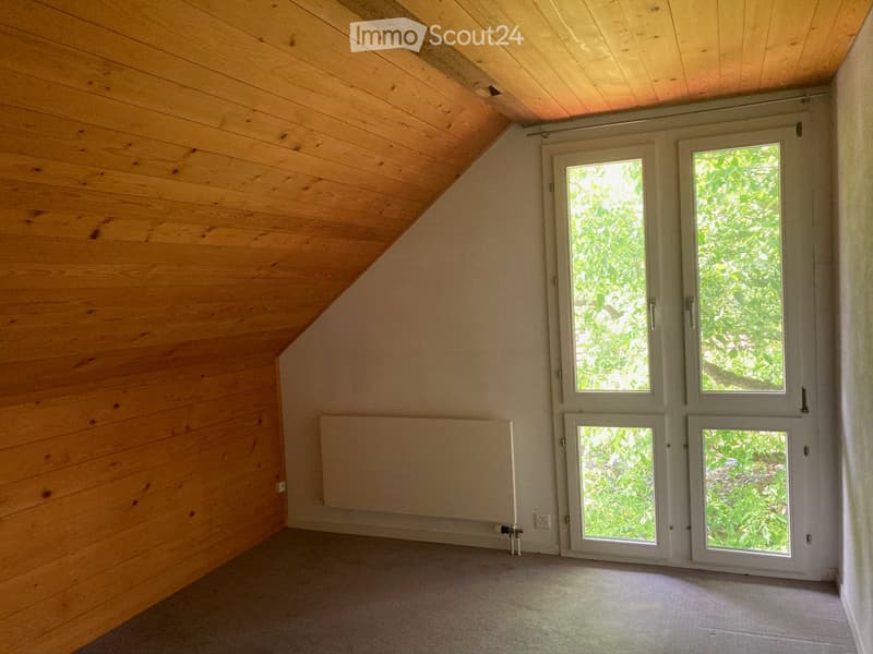 Empty attic room with wooden walls and ceiling, large windows, and a radiator. No furniture or equipment present.