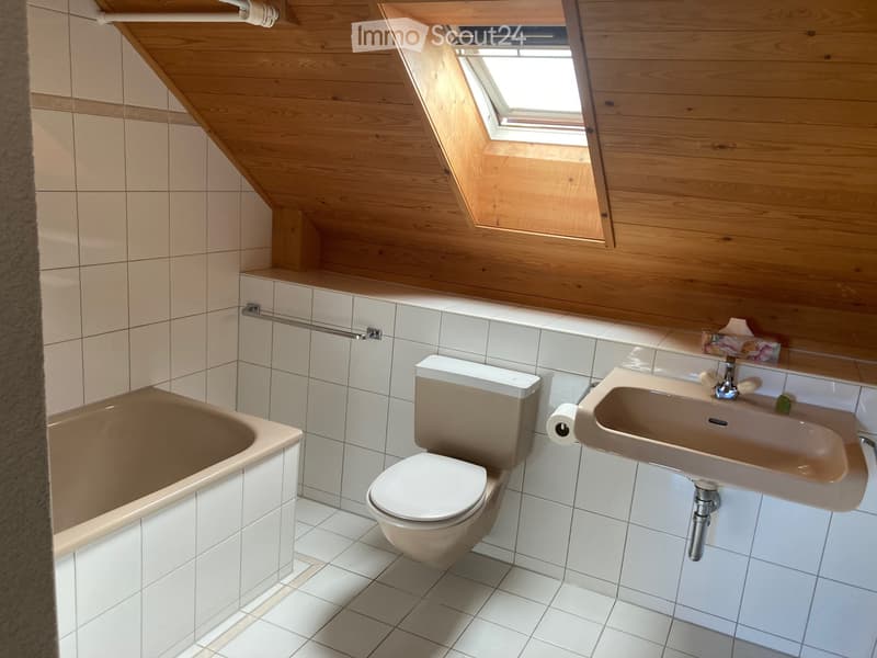 Bathroom with a bathtub, toilet, sink, and a towel rack. The room features white tiled walls and a wooden slanted ceiling with a skylight window.