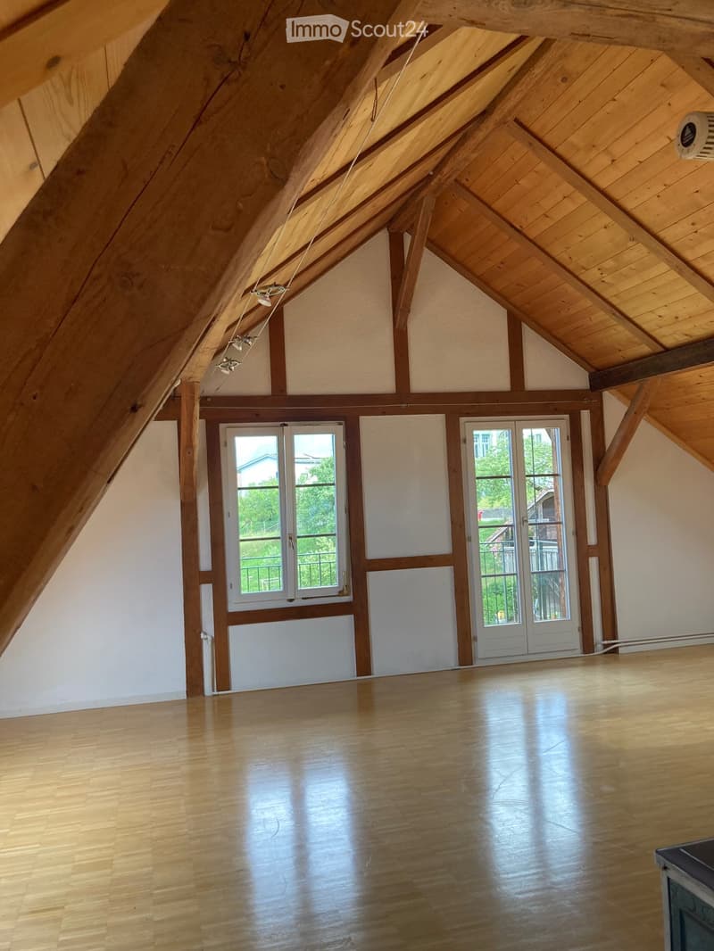 An empty attic space with wooden beams, wooden flooring, and large windows offering natural light. The room features exposed wooden beams and a sloped ceiling.