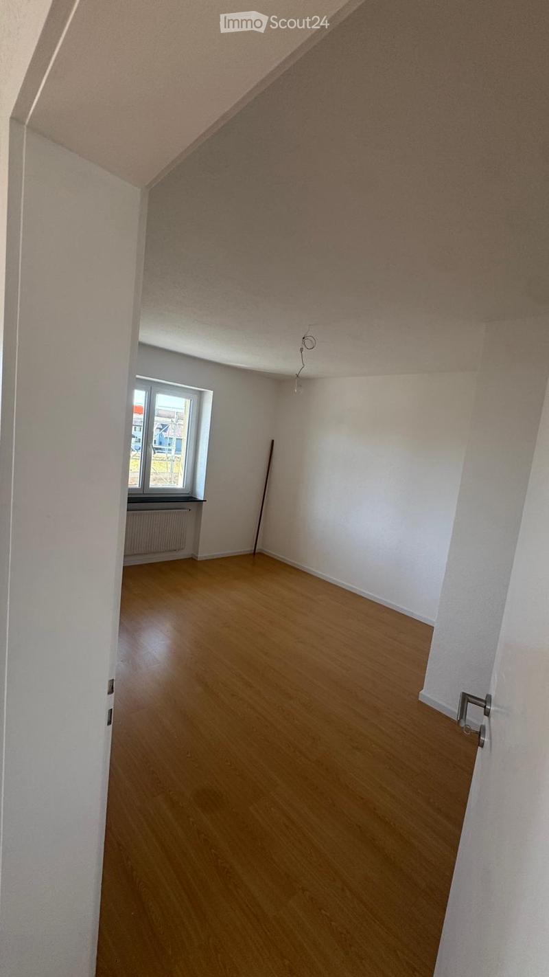 Empty room with wooden flooring, white walls, and a window with a radiator below it. The room has a slanted ceiling and a door visible on the right side.