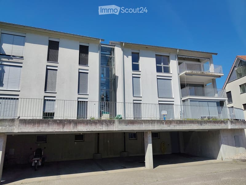 Modern multi-story residential building with white facade, large windows, balconies, and a covered parking area below.