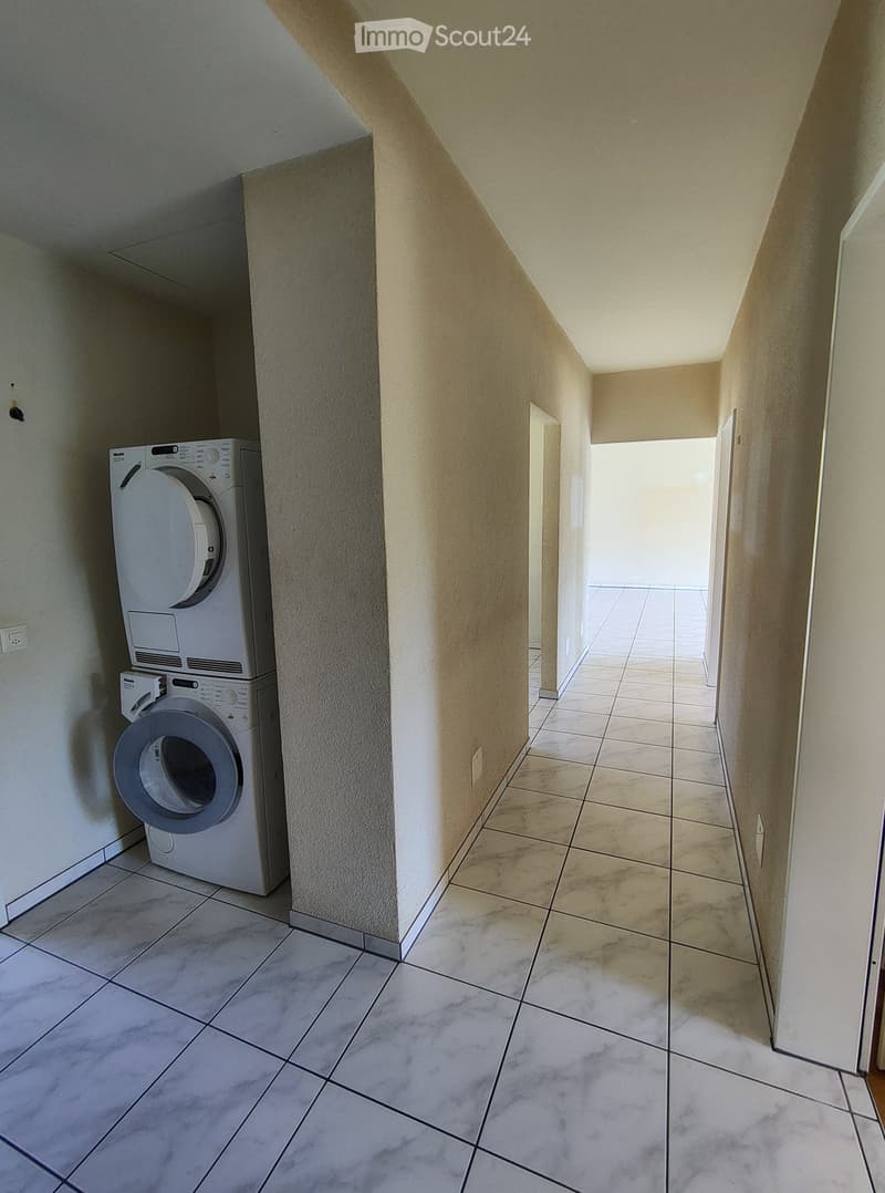 A narrow hallway with tiled flooring and beige walls, featuring a washing machine and dryer unit on the left side.