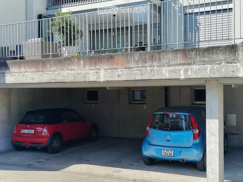 Underground garage with two parked cars, a red and a blue, under a concrete structure with a balcony above.
