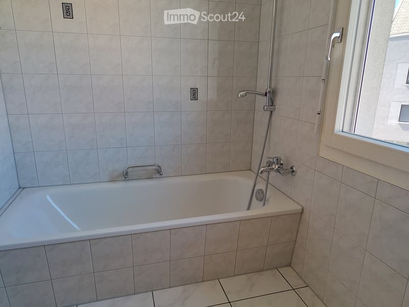 Modern bathroom with a bathtub, tiled walls, and a window. Features a handheld showerhead and a chrome faucet.