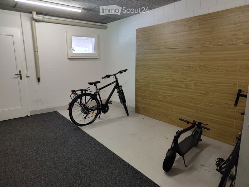 Empty room with a wooden panel wall, a small window, a grey carpet, and two bicycles (one regular and one electric scooter) placed inside.