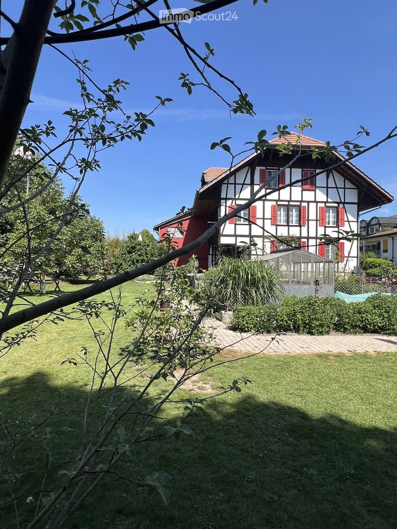 A well-maintained garden with a lawn, trees, and a pathway leading to a house with red shutters and a tiled roof.