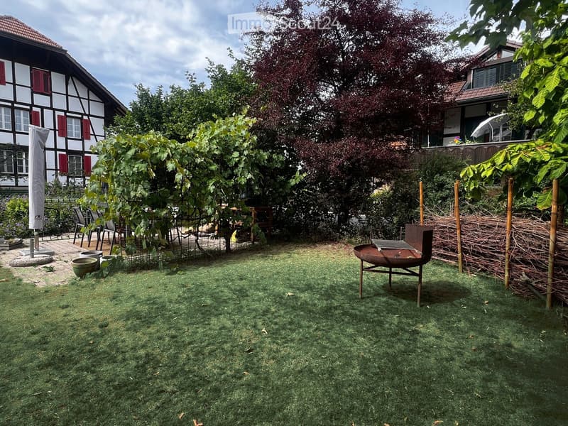 Well-maintained garden with a grassy lawn, barbecue, outdoor furniture, and surrounding trees and shrubs. The background shows a traditional half-timbered house with red shutters and a modern extension.
