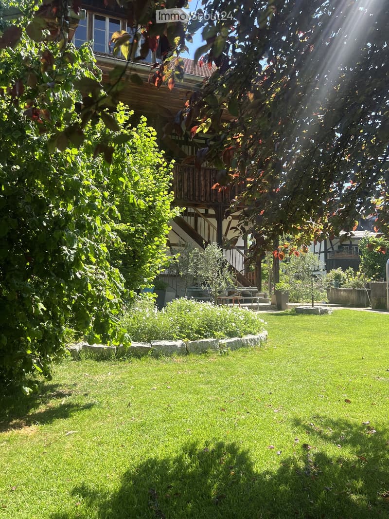 Well-maintained garden with a lush green lawn, surrounded by trees and shrubs, with a wooden deck and staircase leading to a house in the background.