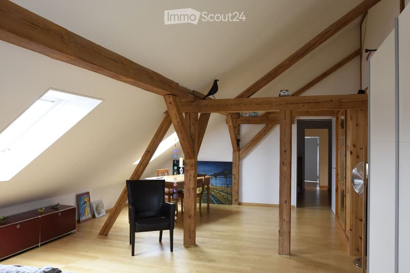 Loft-style living space with wooden beams, skylights, a black leather chair, a dining table with chairs, a red cabinet, and a fan. The room features exposed wooden beams and a high, slanted ceiling.