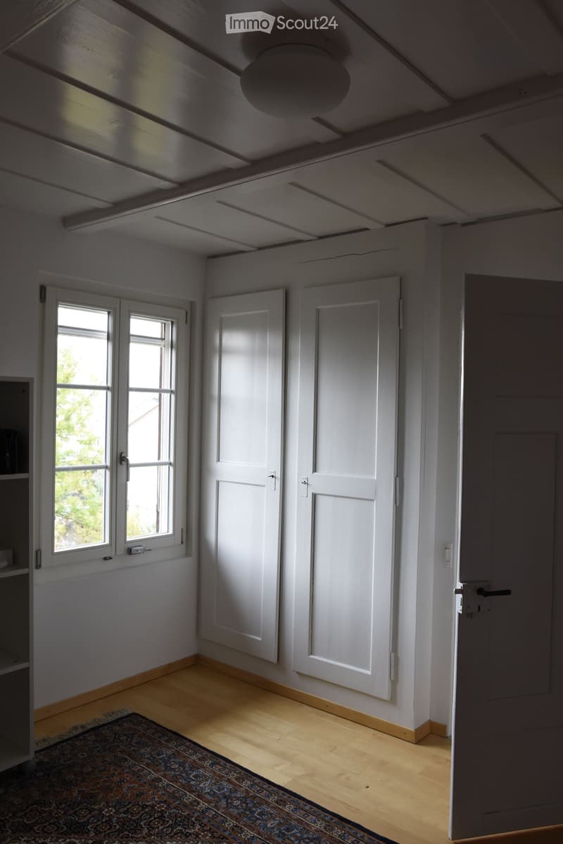 Empty room with wooden flooring, white walls, and white doors. A small window on the left side allows natural light to enter. A patterned rug is placed on the floor near the center of the room.
