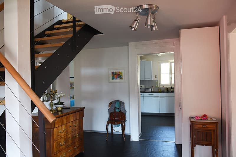 Hallway with a wooden staircase, a small wooden table with a vase, a framed picture on the wall, and a view into a kitchen area with white cabinets and a window.