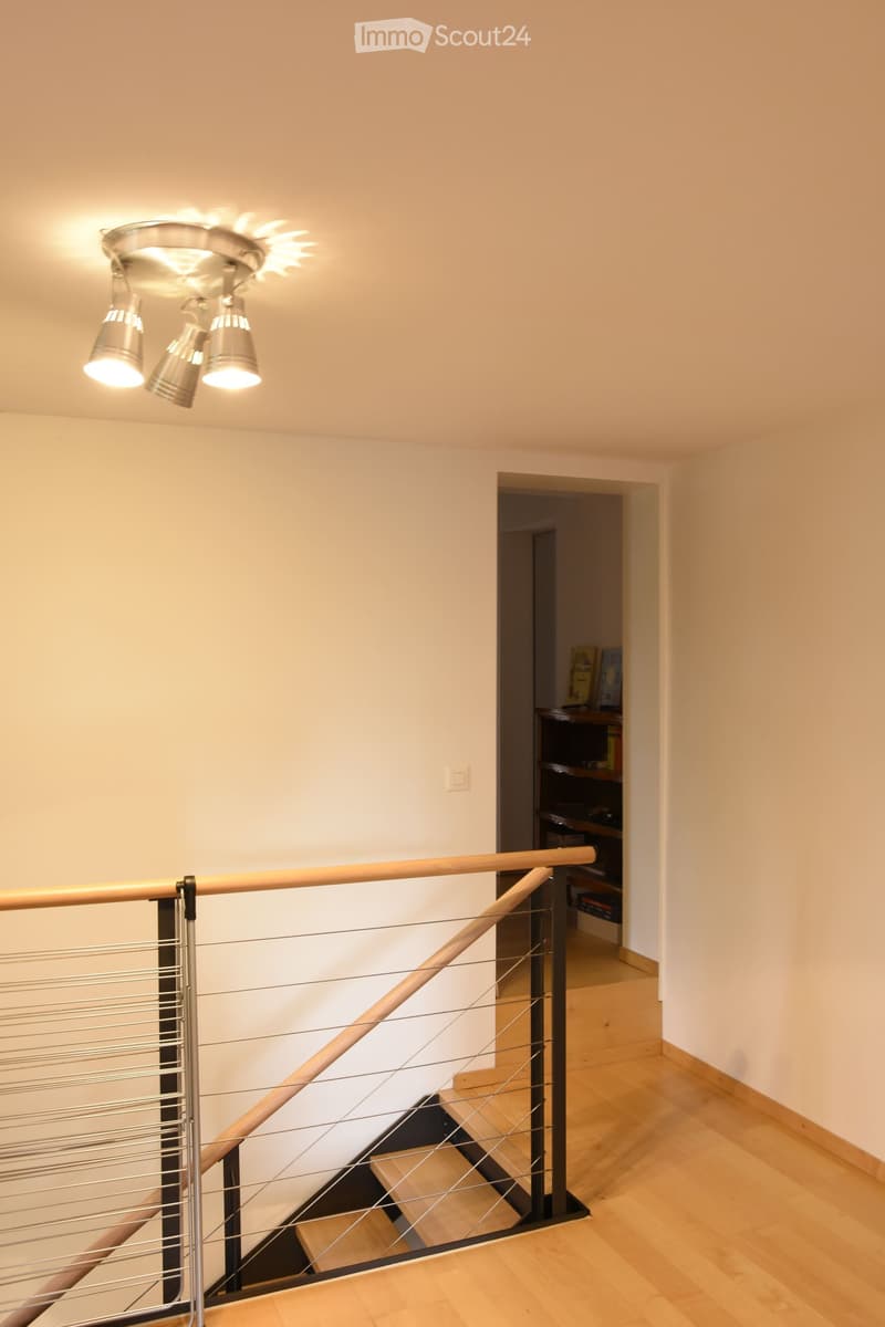 Modern hallway with wooden flooring, a staircase featuring a metal railing and wooden handrail, and a ceiling light fixture with multiple bulbs. A doorway leads to another room with visible furniture.