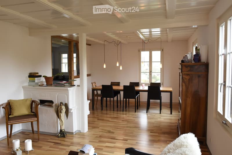 Bright, open living and dining area with wooden floors, white walls, and exposed wooden beams. Features a fireplace with a decorative angel statue, a dining table with six chairs, a wooden cabinet, and a cozy armchair with a green pillow.